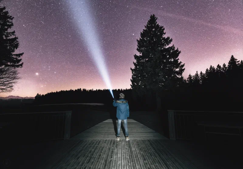 Person in a blue jacket on a wooden bridge shining a flashlight at the night sky
